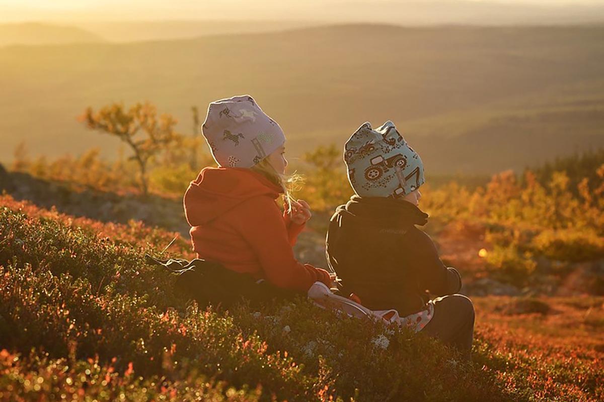 Två barn sitter i höstfärgad fjällterräng och tittar ut över solnedgången